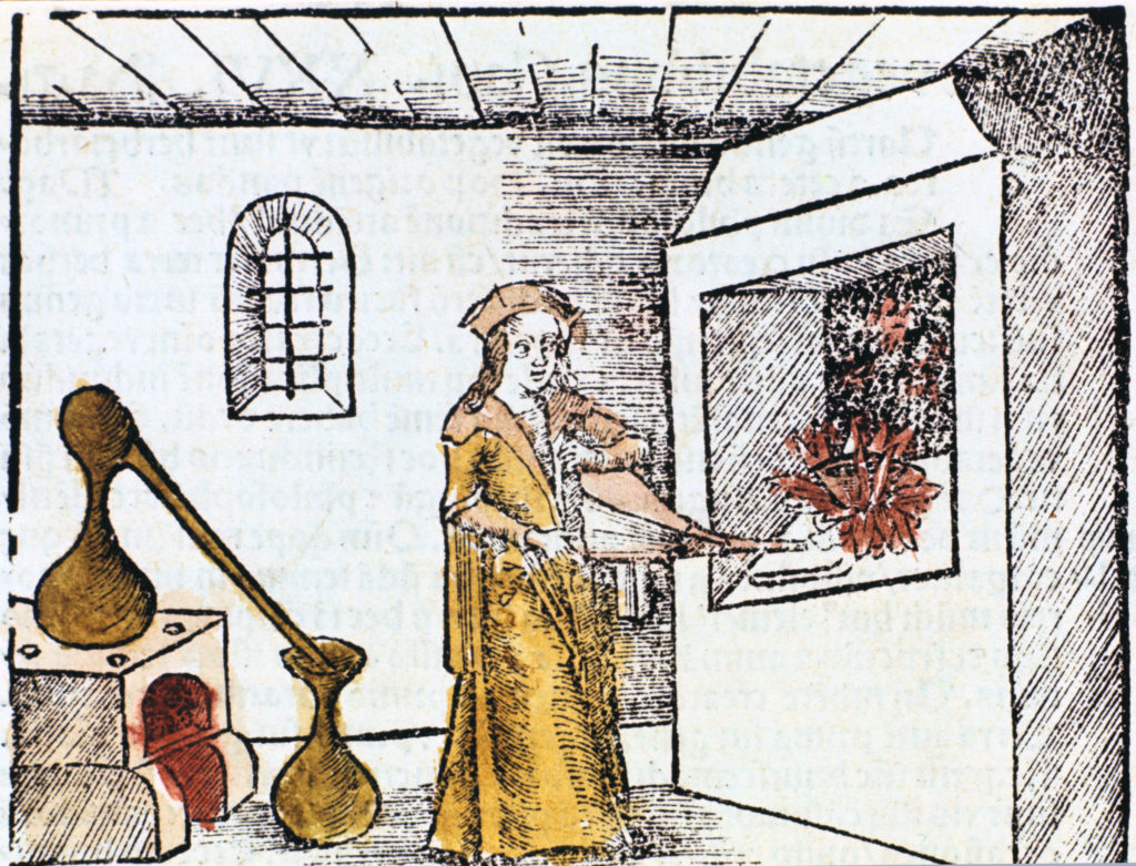 A chemist using a bellows to encourage a fire burning as part of an experiment. Various vessels and equipment are shown elsewhere in the room.